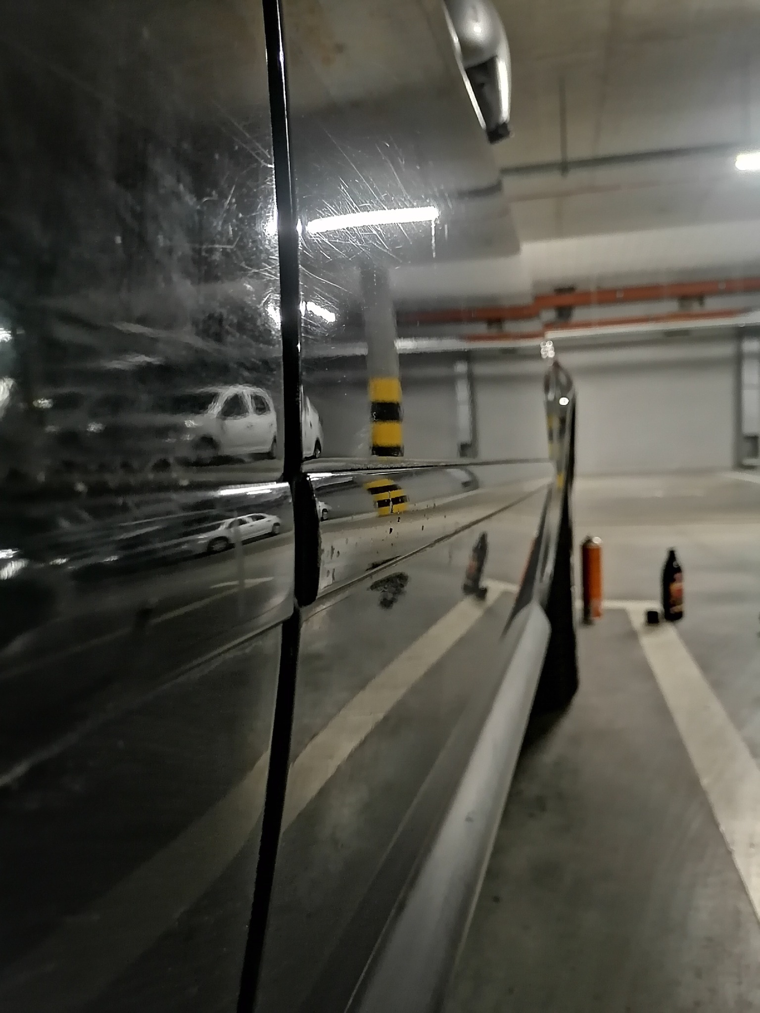 Błyszcząca, czarna karoseria samochodu z widocznymi zarysowaniami i odbiciami innych aut na parkingu podziemnym, obok stoją dwie butelki z preparatami do pielęgnacji auta.