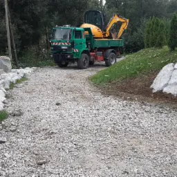 Zielona ciężarówka z żółtą mini-koparką na platformie, zaparkowana na żwirowej drodze otoczonej dużymi kamieniami i zielenią.