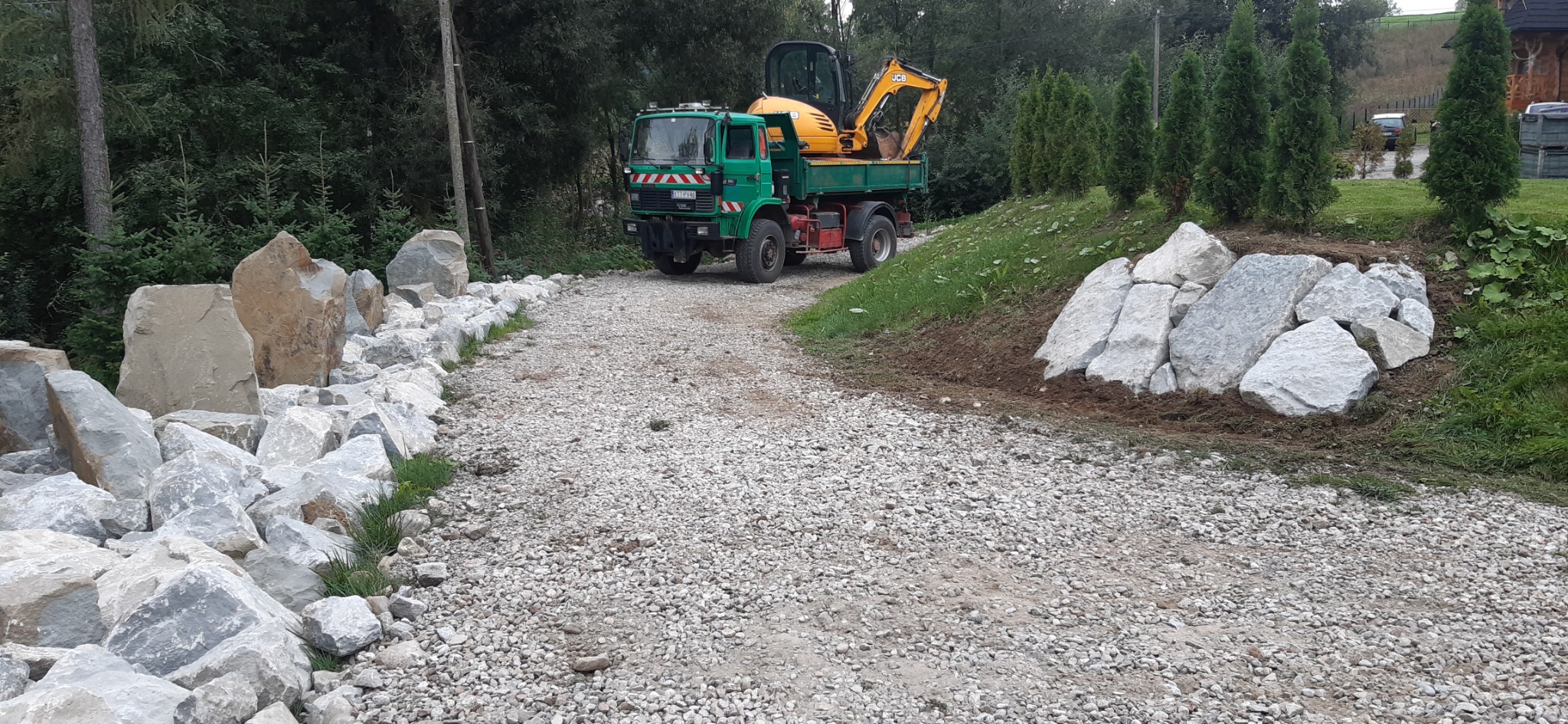 Zielona ciężarówka z żółtą mini-koparką na platformie, zaparkowana na żwirowej drodze otoczonej dużymi kamieniami i zielenią.