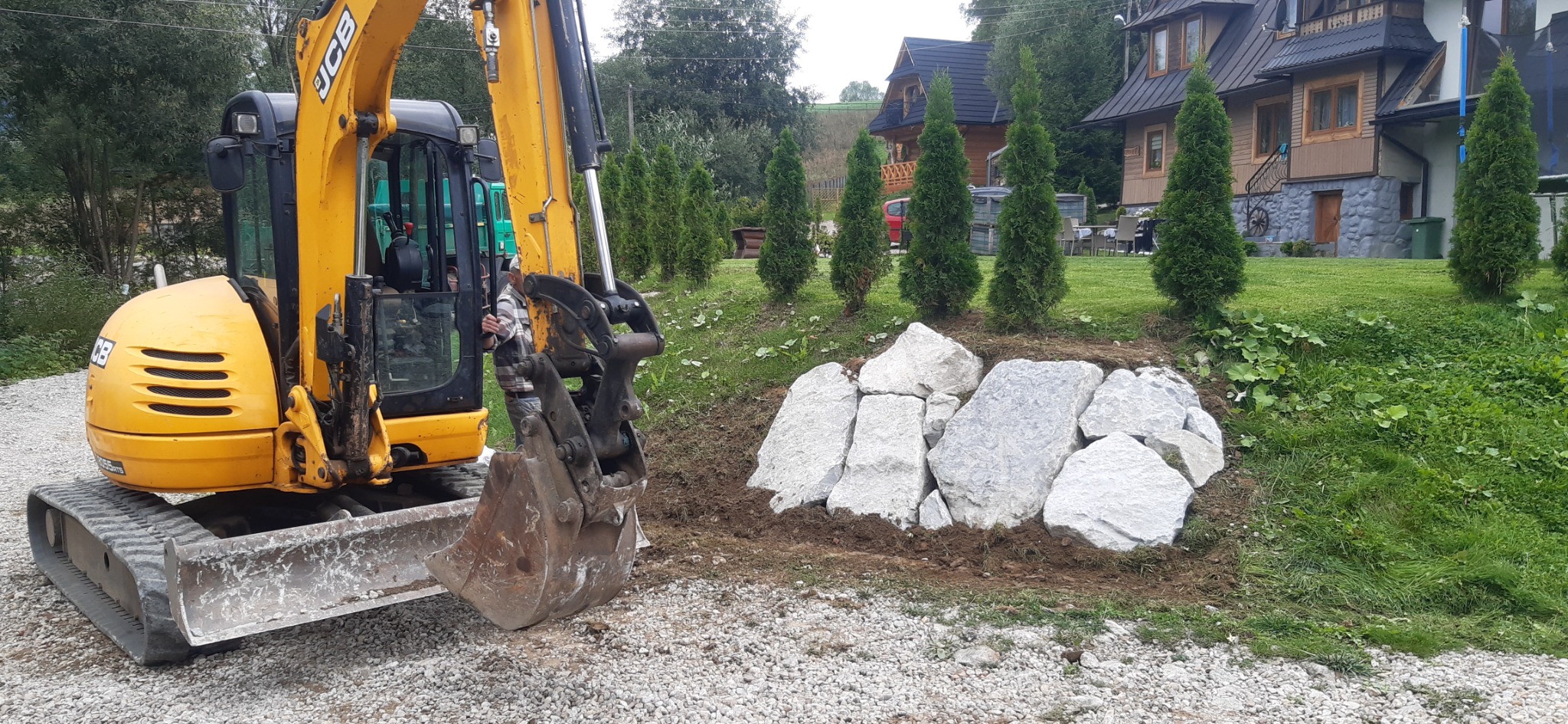 Żółta koparka JCB z łyżką w trakcie prac ziemnych przy układaniu dużych, jasnych kamieni na tle zielonego trawnika i domów w stylu góralskim.