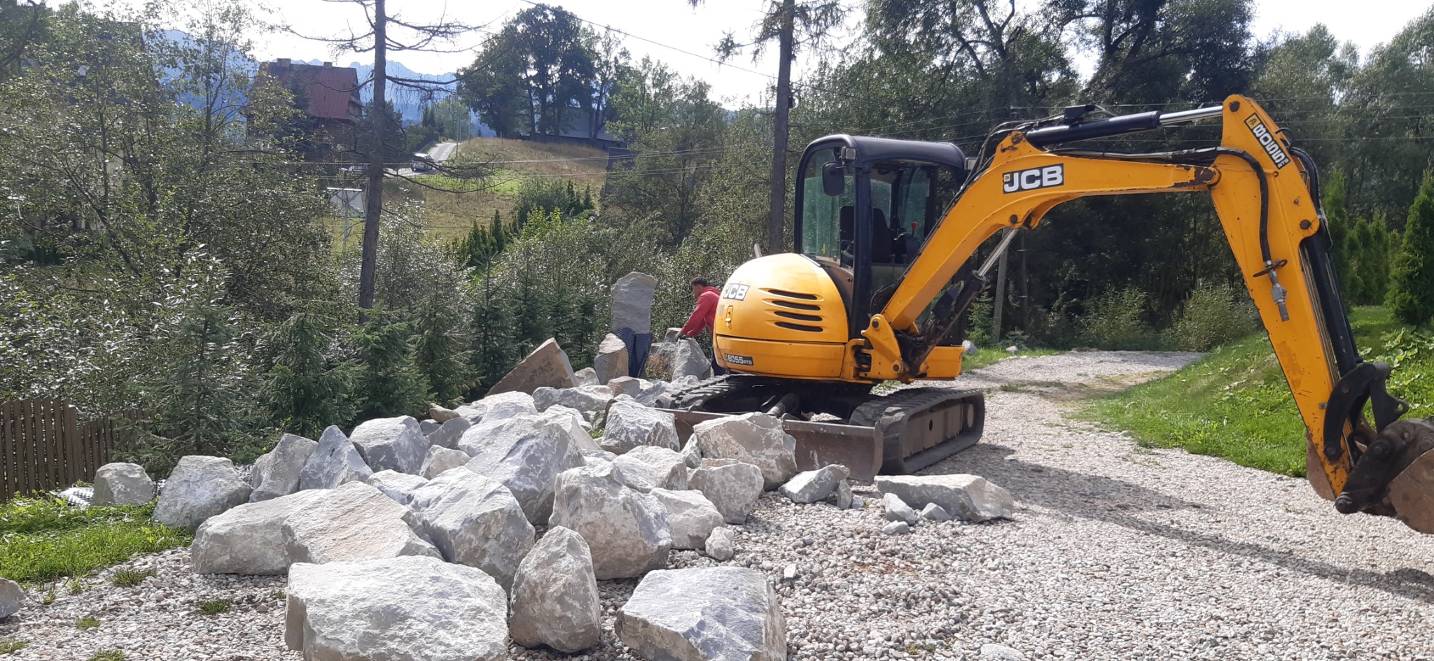 Żółta koparka JCB 8055 ZTS na żwirowym podłożu, obok sterta dużych, jasnych kamieni, w tle zielone drzewa i budynki w górskim krajobrazie, osoba w czerwonej bluzie przy kamieniach.