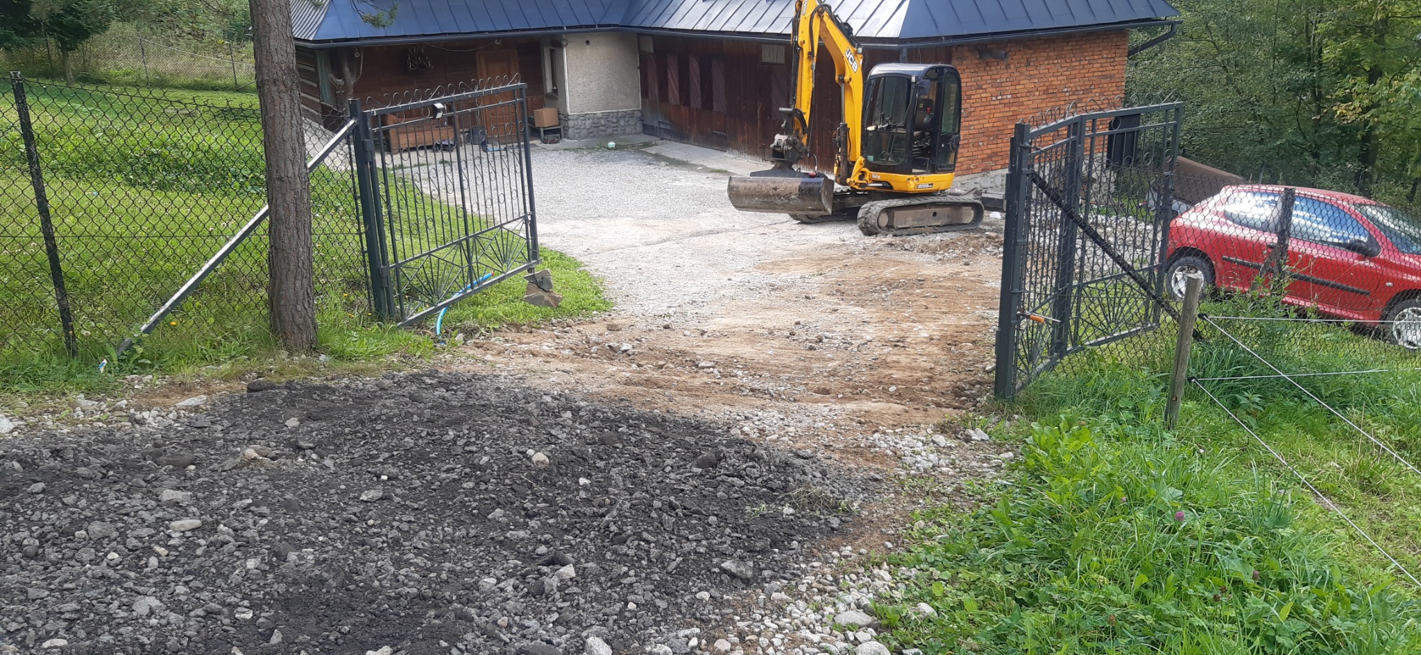 Żółta minikoparka na posesji z otwartą bramą wjazdową, świeżo naruszona ziemia, w tle dom z cegły i czerwony samochód za ogrodzeniem.