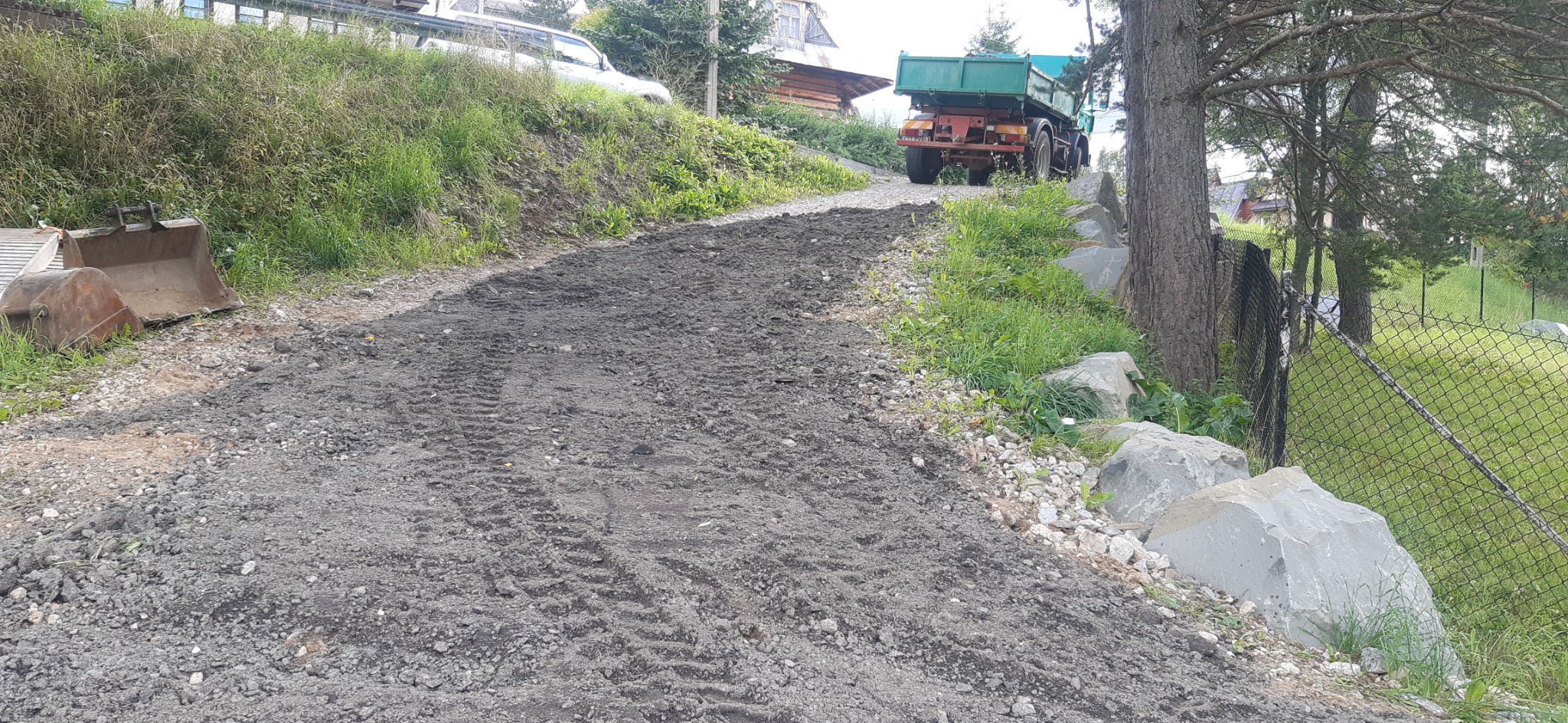 Przygotowanie terenu pod budowę drogi dojazdowej na zboczu, widoczne ślady opon ciężarówki i łyżka koparki obok.