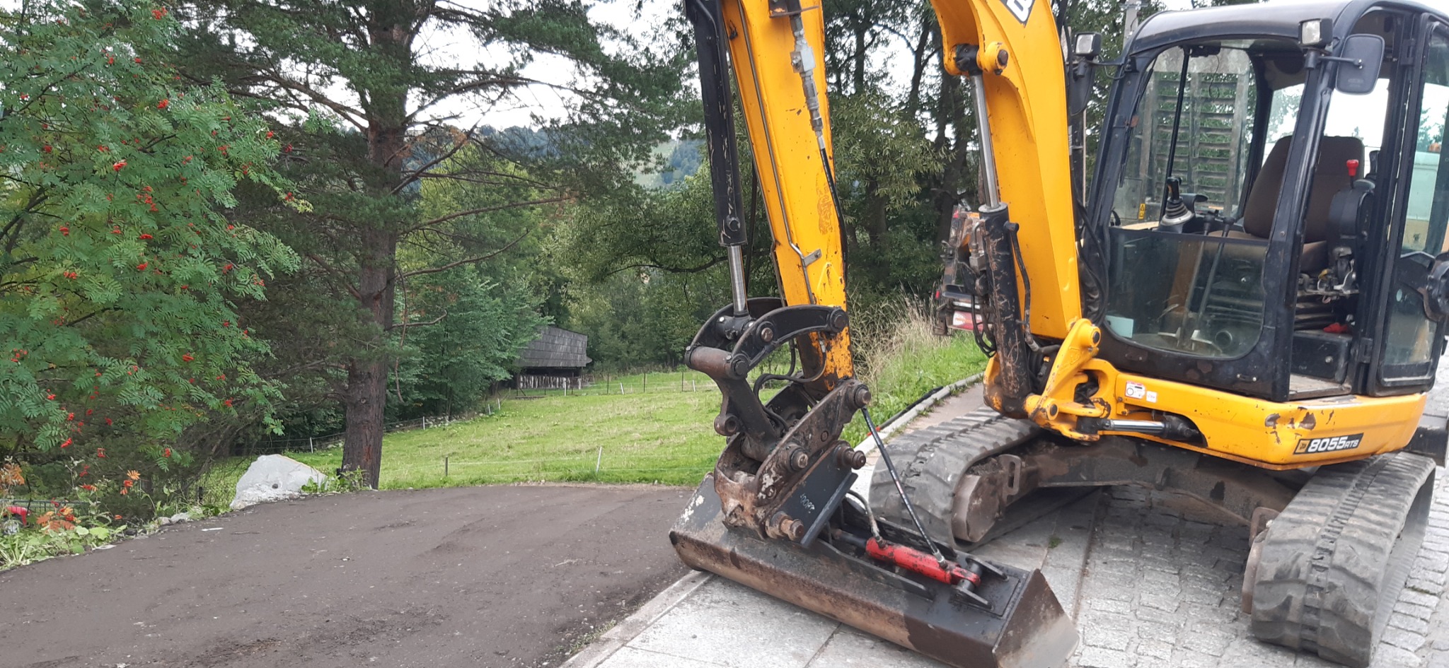 Żółta minikoparka JCB 8055RTB na brukowanej powierzchni, z widocznym wykopem ziemnym w tle i malowniczym krajobrazem górskim zalesionym drzewami liściastymi i iglastymi oraz drewnianą chatą.