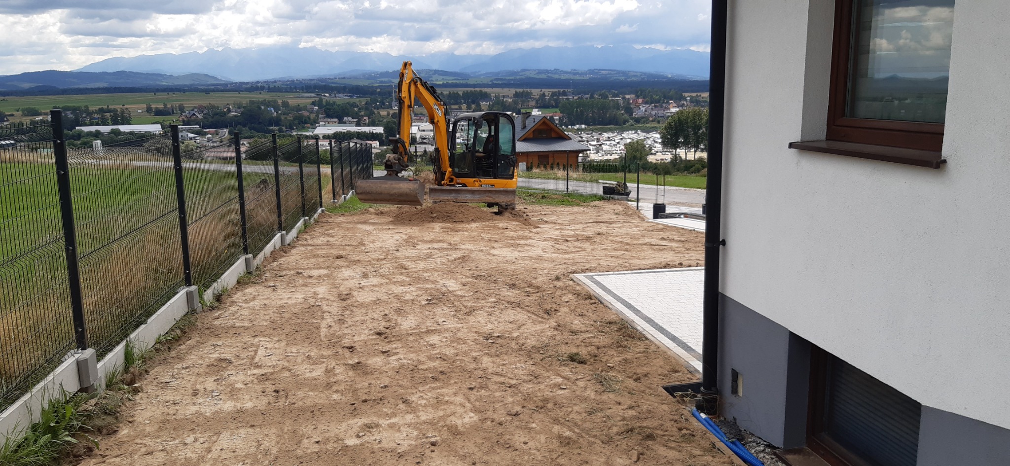 Żółta mini koparka na placu budowy z widokiem na góry w tle, przygotowany teren pod budowę z widocznym fragmentem kostki brukowej i ogrodzeniem.