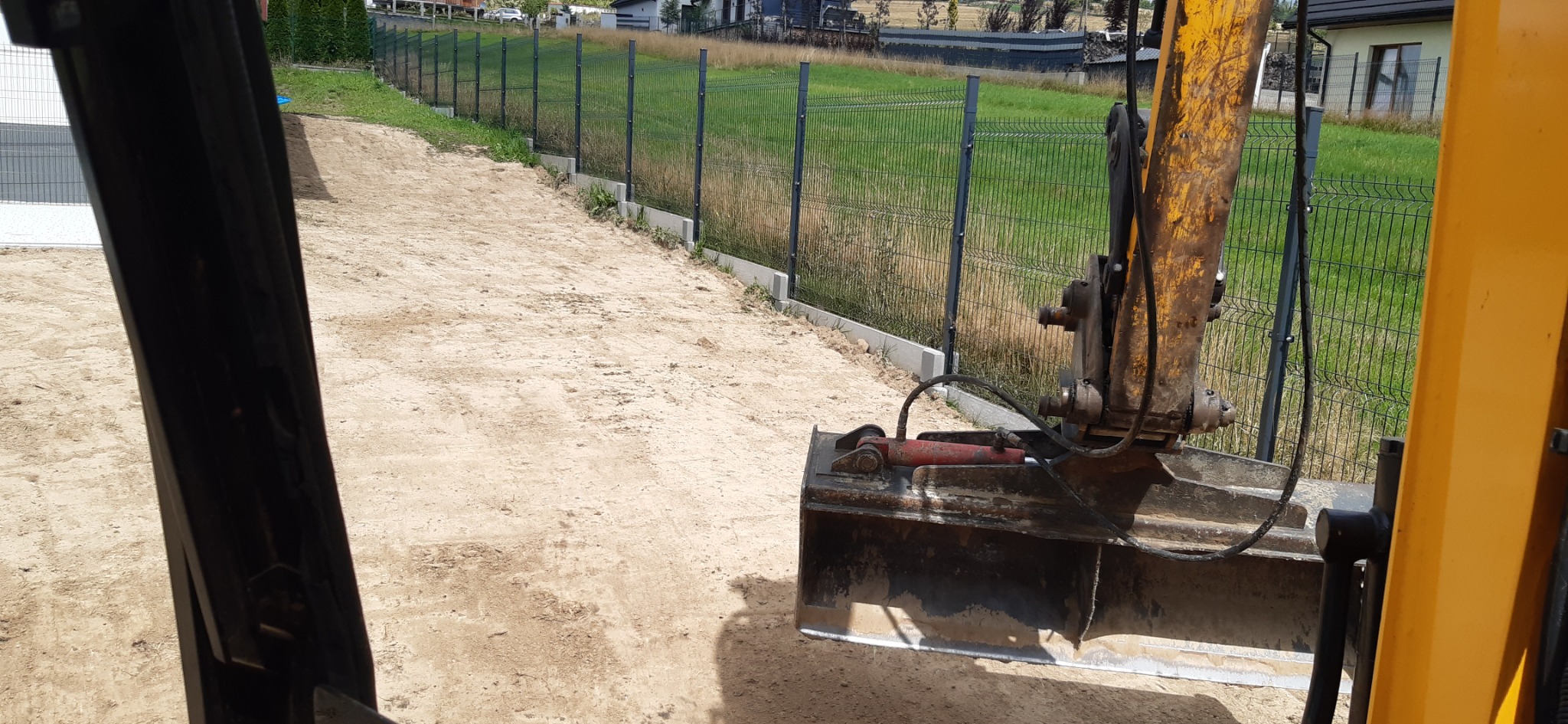 Żółta koparka z łyżką w trakcie prac ziemnych przy ogrodzeniu panelowym na podbudowie betonowej, widok z kabiny operatora.