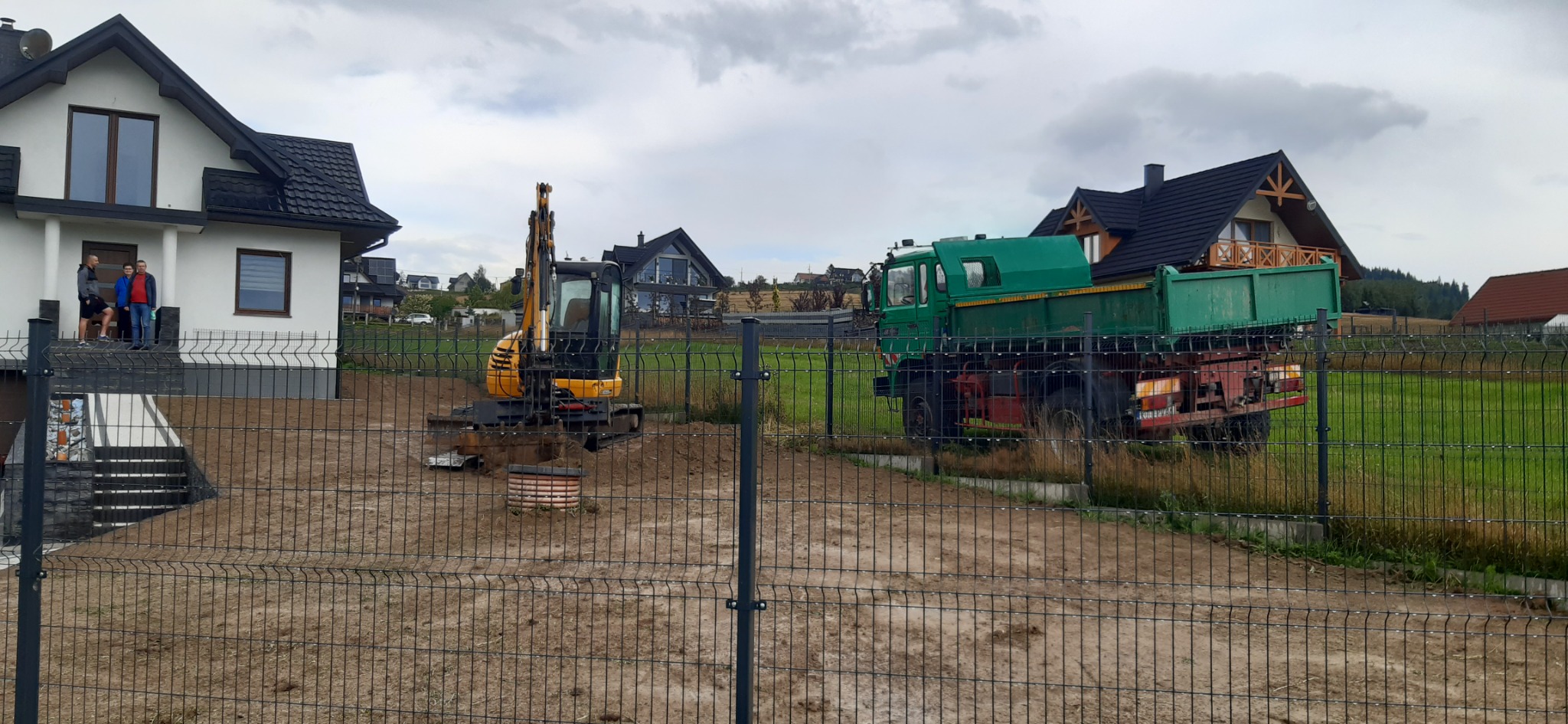 Mini koparka żółto-czarna podczas prac ziemnych na działce z nowo wybudowanym domem, widoczny wykop i zielona ciężarówka w tle, trzech mężczyzn obserwuje prace z ganku.