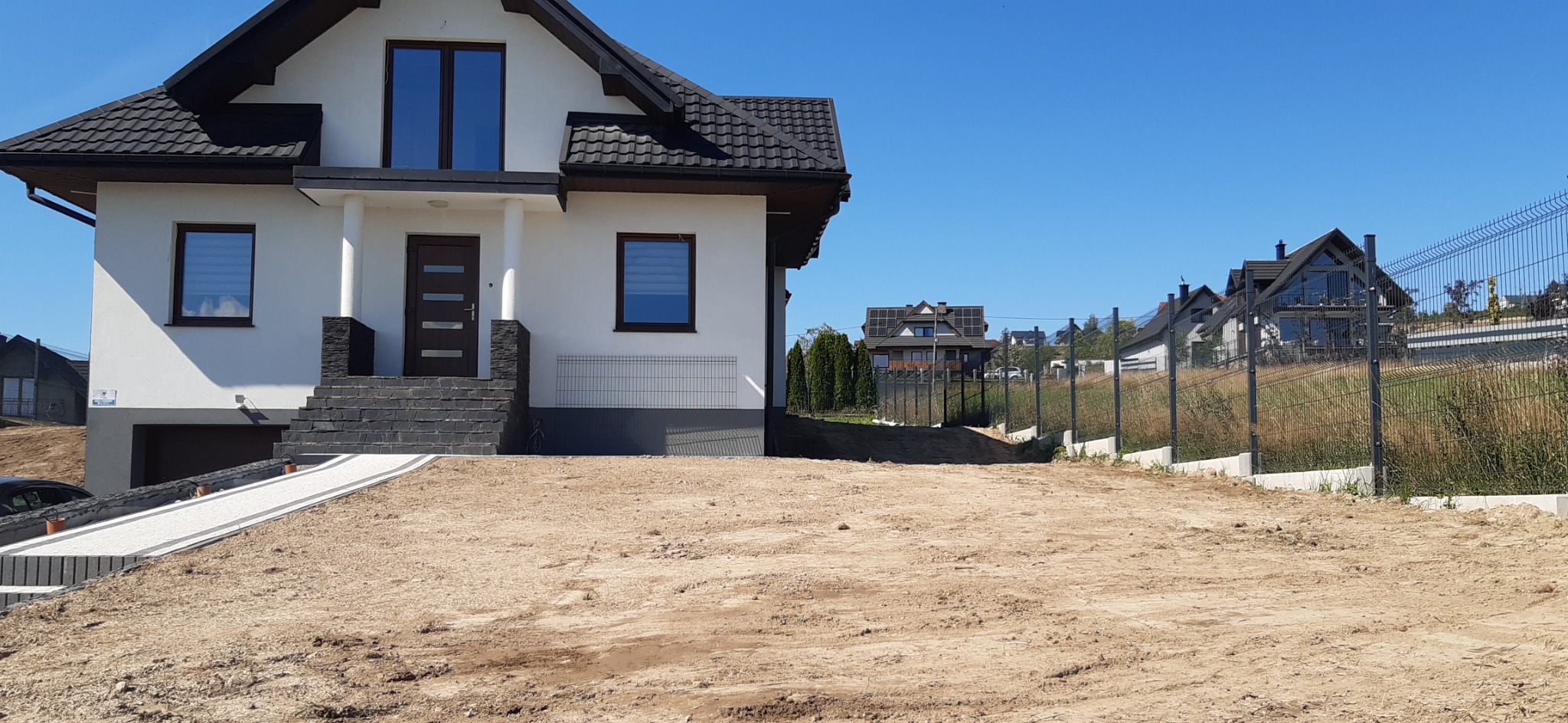 Nowoczesny dom jednorodzinny z czarną dachówką i ciemnymi oknami, widok na teren przygotowany pod ogród i ogrodzenie panelowe.