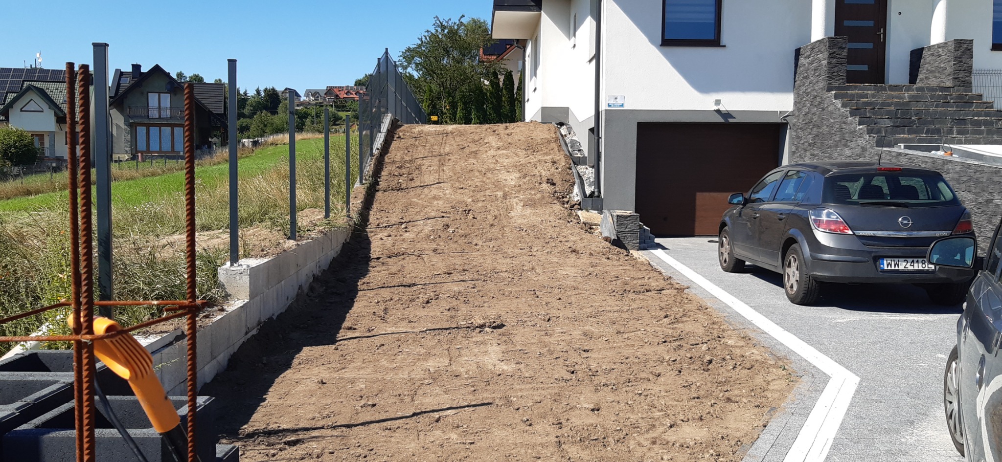 Przygotowany podjazd z ziemi do posesji z widocznymi fundamentami pod ogrodzenie i zaparkowanym samochodem osobowym.