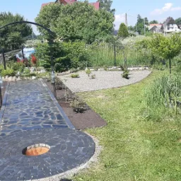 Ogr&oacute;d z ścieżką z nieregularnych kamieni prowadzącą do okrągłego paleniska, nowo posadzone krzewy r&oacute;ż wzdłuż ścieżki, żwirowy obszar z młodymi drzewkami, całość otoczona zielenią i ogrodzeniem.