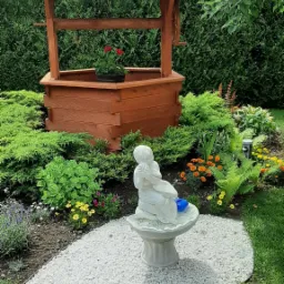 Ogród z drewnianą studnią, kwiatami, krzewami i białą figurką dziecka na postumencie otoczonym białym żwirem.