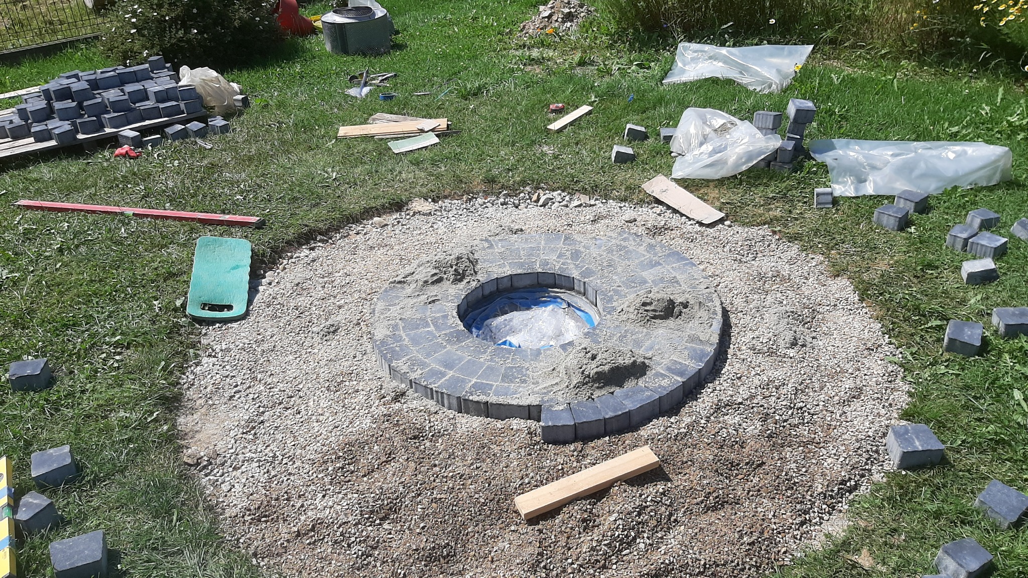Budowa okrągłego paleniska z szarej kostki brukowej na podsypce żwirowej, widoczne narzędzia i materiały budowlane na trawie.