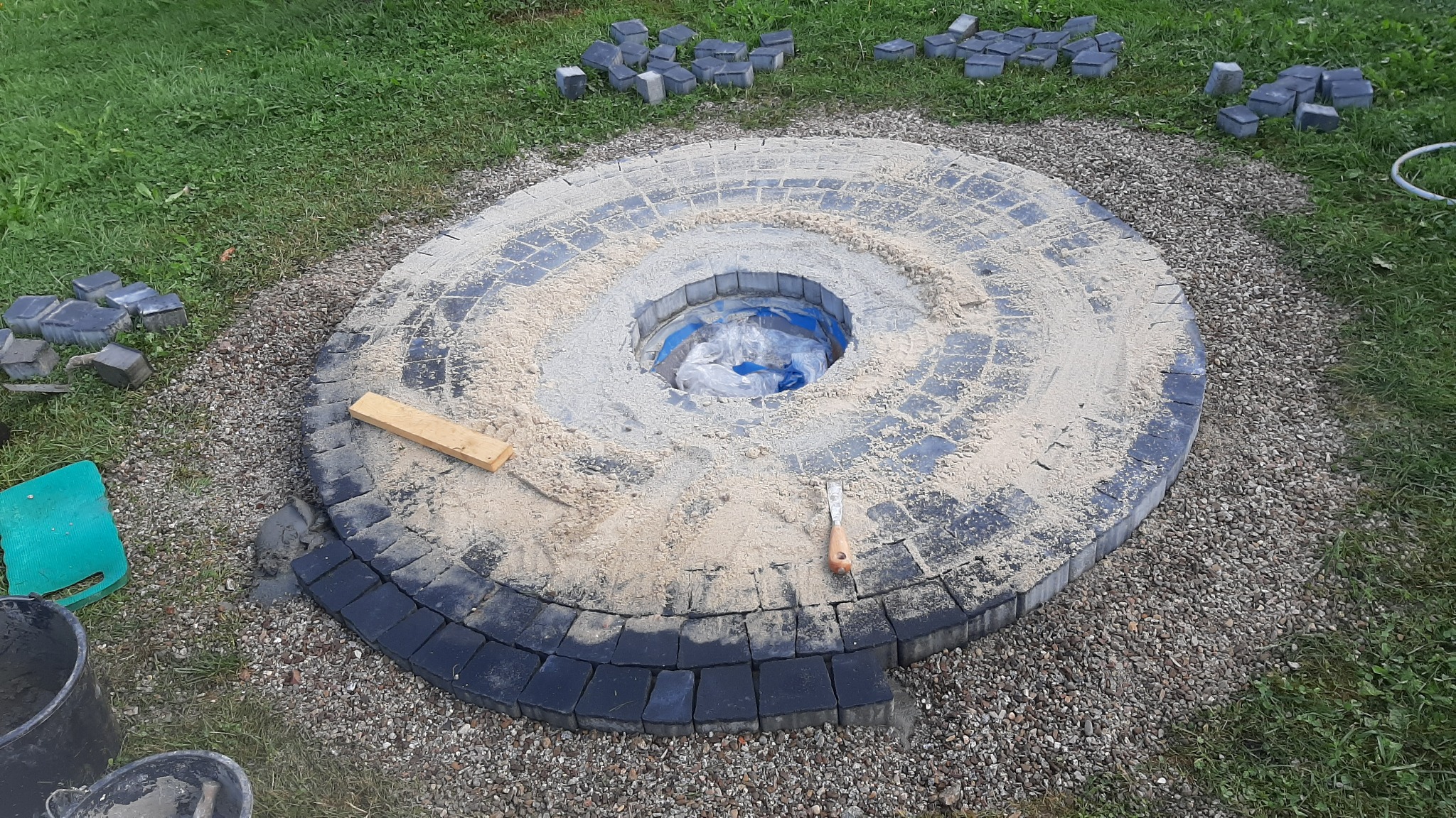 Budowa okrągłego paleniska z czarnej kostki brukowej, częściowo zasypanego piaskiem, z widocznymi narzędziami i zapasem kostki na trawie.