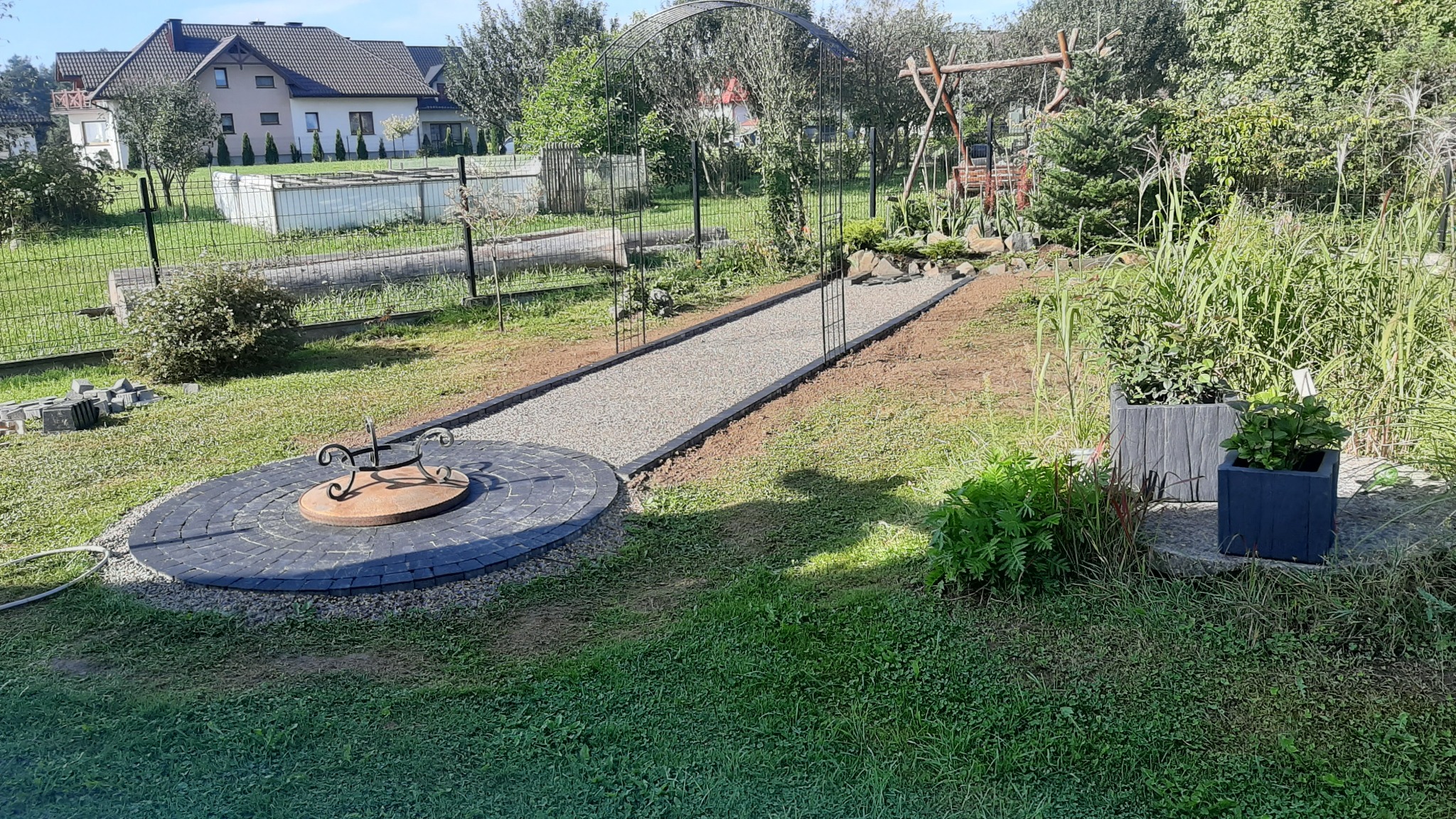 Ogród z wytyczoną ścieżką żwirową, okrągłym paleniskiem z kostki brukowej i donicami z roślinami, w tle huśtawka i domy.
