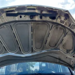 Otwarta maska samochodu z widocznymi śladami zabrudzeń i zużycia, uchwyt podtrzymujący maskę po lewej stronie, na tle muru z cegły i fragmentu nieba.