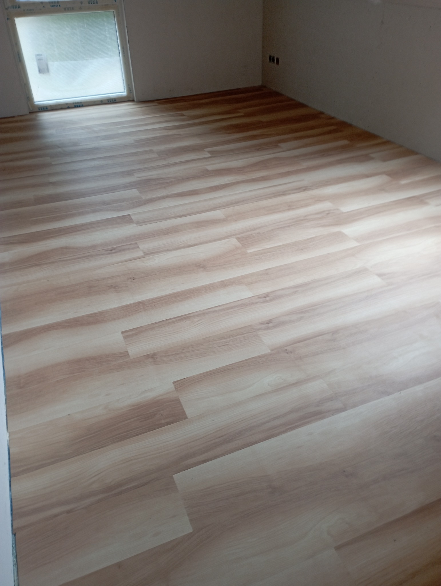 Jasne panele podłogowe imitujące drewno ułożone w pokoju z oknem, widoczne gniazdka elektryczne na ścianie.