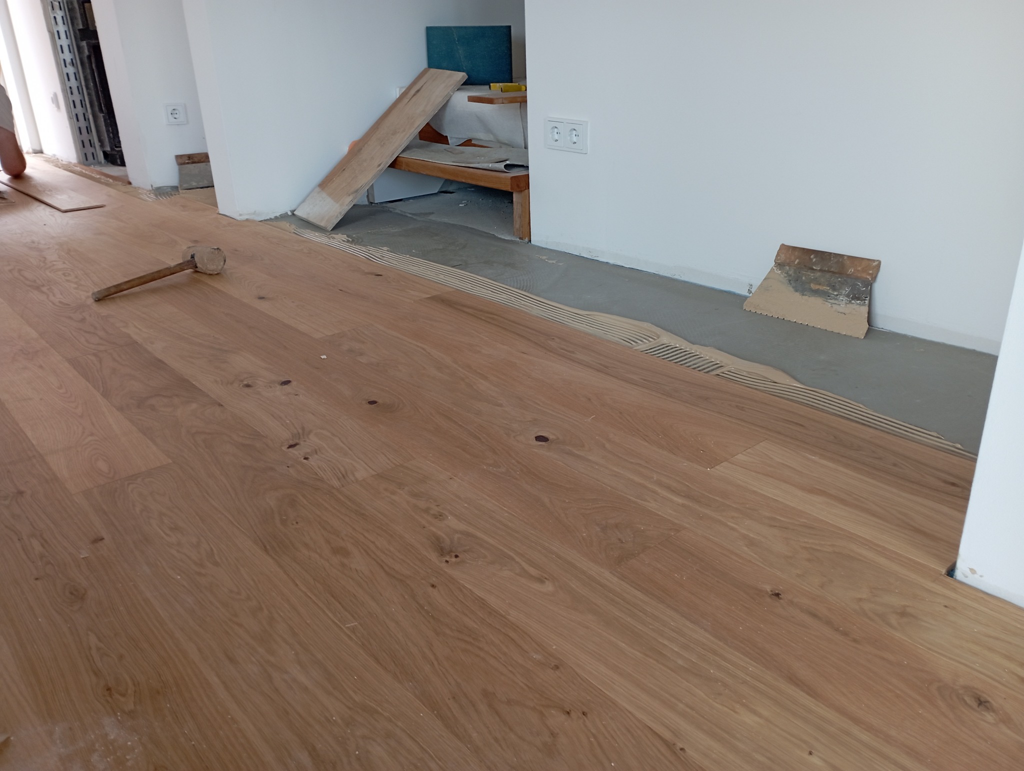 Fragment podłogi z klejem i częściowo ułożonym parkietem, widoczny gumowy młotek i szpachelka przy ścianie.