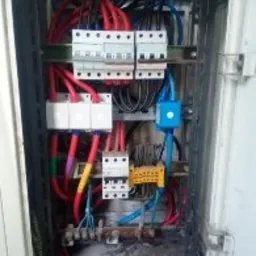 Otwarta skrzynka elektryczna z widocznymi bezpiecznikami, przewodami w izolacji czerwonej, niebieskiej i żółtej, oraz listwami zaciskowymi. Widoczne ślady korozji na elementach metalowych...