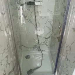 Kabina prysznicowa z brodzikiem i chromowaną armaturą, wykończona panelami ściennymi imitującymi marmur, widoczna szpachelka na dnie brodzika.