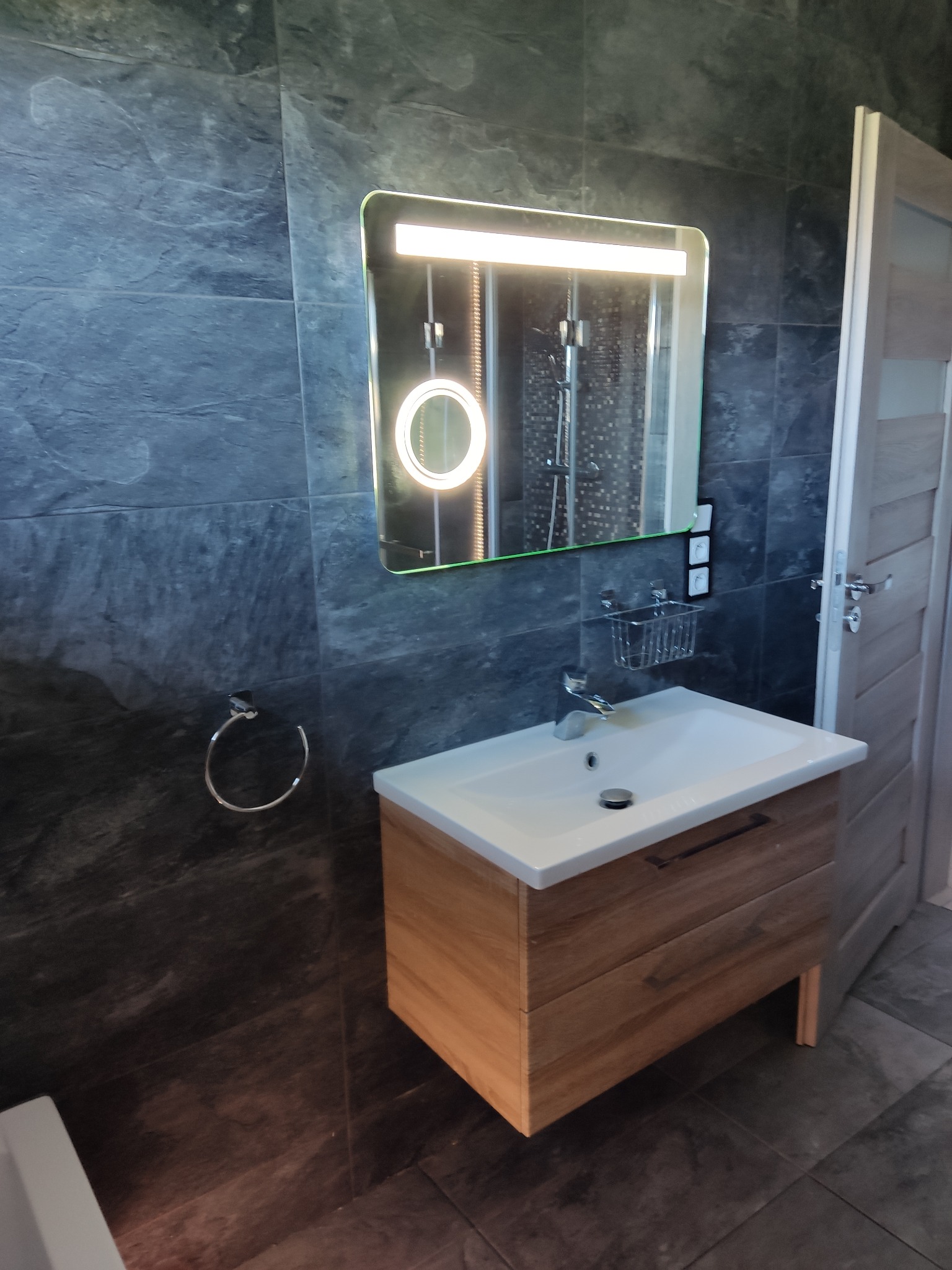 Nowoczesna łazienka z umywalką na drewnianej szafce, podświetlanym lustrem i ciemnymi płytkami imitującymi kamień. W tle widoczna kabina prysznicowa i drzwi.