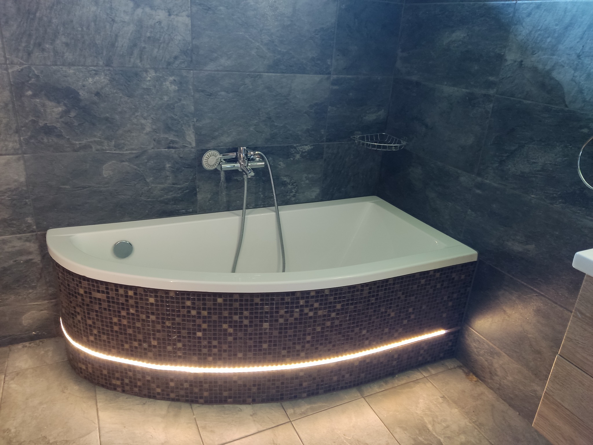 Nowoczesna wanna narożna obudowana mozaiką z podświetleniem LED, szara ściana z płytek imitujących kamień, chromowana bateria i półka w łazience.