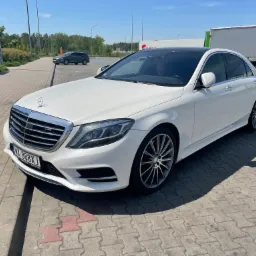 Biały Mercedes-Benz S-Class z czarnym dachem panoramicznym, zaparkowany na parkingu z kostki brukowej, z widoczną tablicą rejestracyjną i przyciemnionymi szybami.