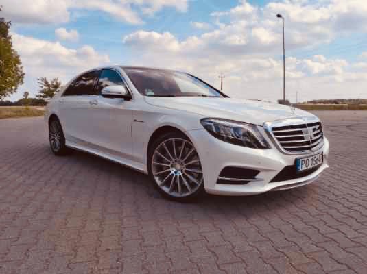 Biały Mercedes-Benz klasy S, widok z boku na tle błękitnego nieba z chmurami, zaparkowany na brukowanej powierzchni.