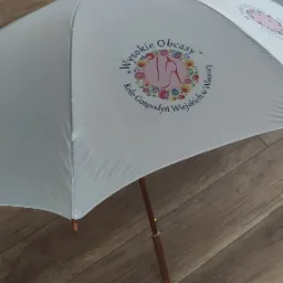Biały parasol z nadrukowanym kolorowym logo 'Wysokie Obcasy' Koło Gospodyń Wiejskich w Winnicy, leżący na drewnianej podłodze.
