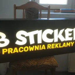 Podświetlany szyld reklamowy z napisem 'DB STICKERS - PRACOWNIA REKLAMY' na czarnym tle, ustawiony na krześle w pomieszczeniu.