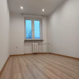 Puste, jasne pomieszczenie po remoncie z panelami podłogowymi, białymi ścianami, oknem i grzejnikiem; widoczne gniazdko elektryczne na ścianie.