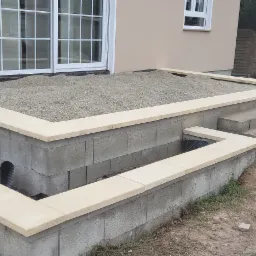 Przygotowany fundament pod taras z szarej kostki betonowej, obłożony kremowymi obrzeżami, widoczne schody, drenaż oraz zasypka z drobnego żwiru, w tle dom jednorodzinny i trampolina.