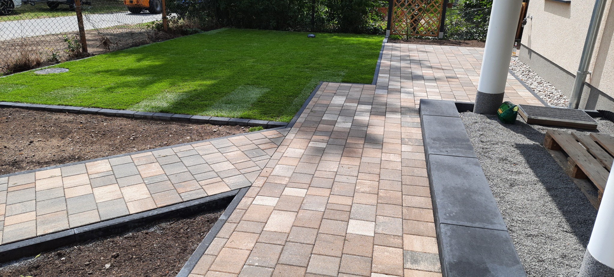 Ułożona ścieżka z kostki brukowej w odcieniach beżu i szarości, obrzeża z ciemnego kamienia, trawnik z rolki, widok na ogród i fragment elewacji budynku.