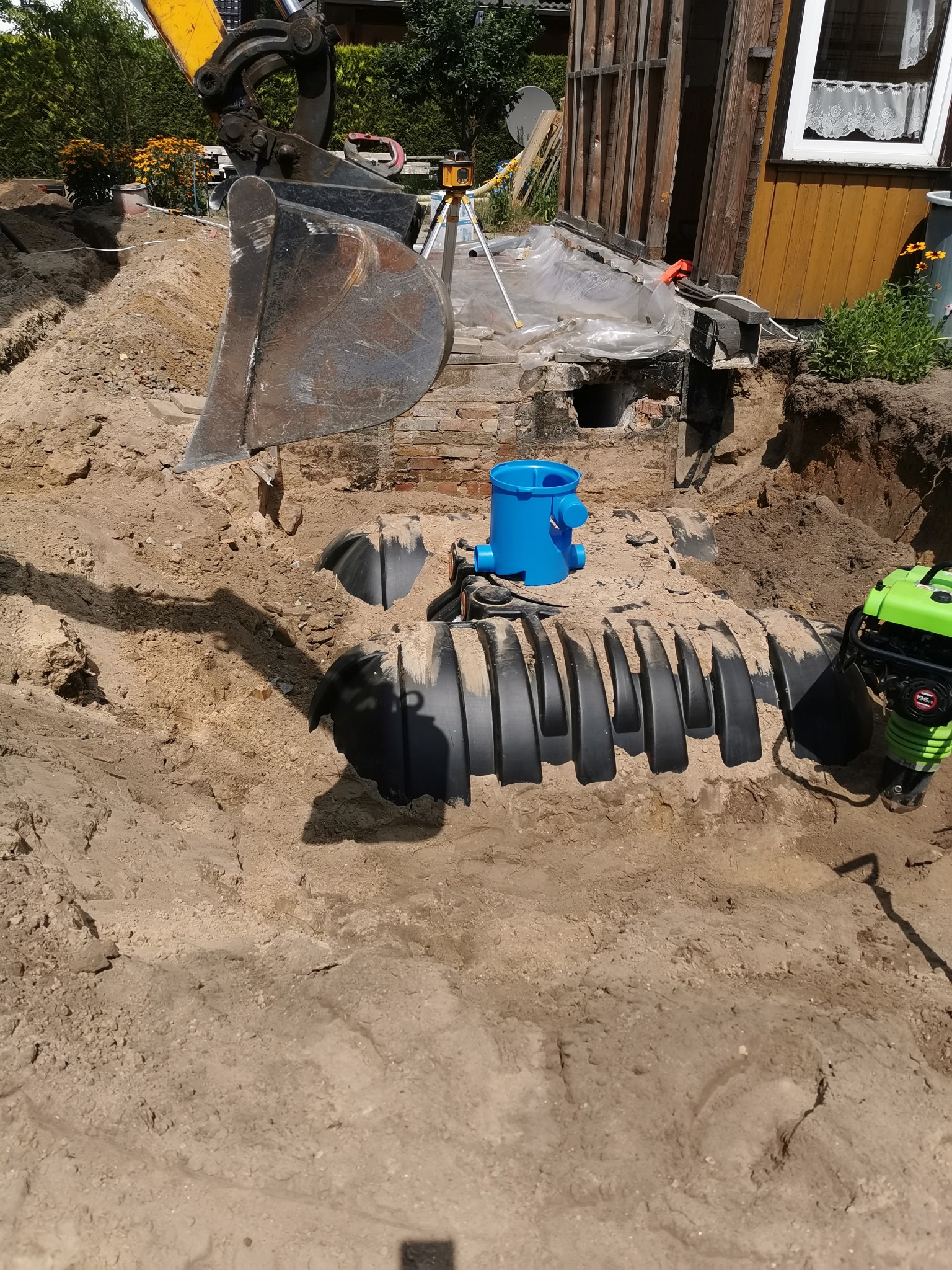 Wykop w ziemi z częściowo wkopanym zbiornikiem z tworzywa sztucznego, niebieski element studzienki rewizyjnej, ubijarka wibracyjna i łyżka koparki w tle, widoczne fundamenty budynku.