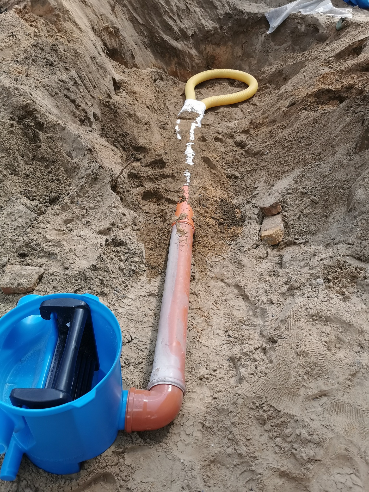 Wykop w ziemi z ułożoną rurą kanalizacyjną, żółtą rurą drenażową i niebieską studzienką rewizyjną z czarnym zamknięciem.