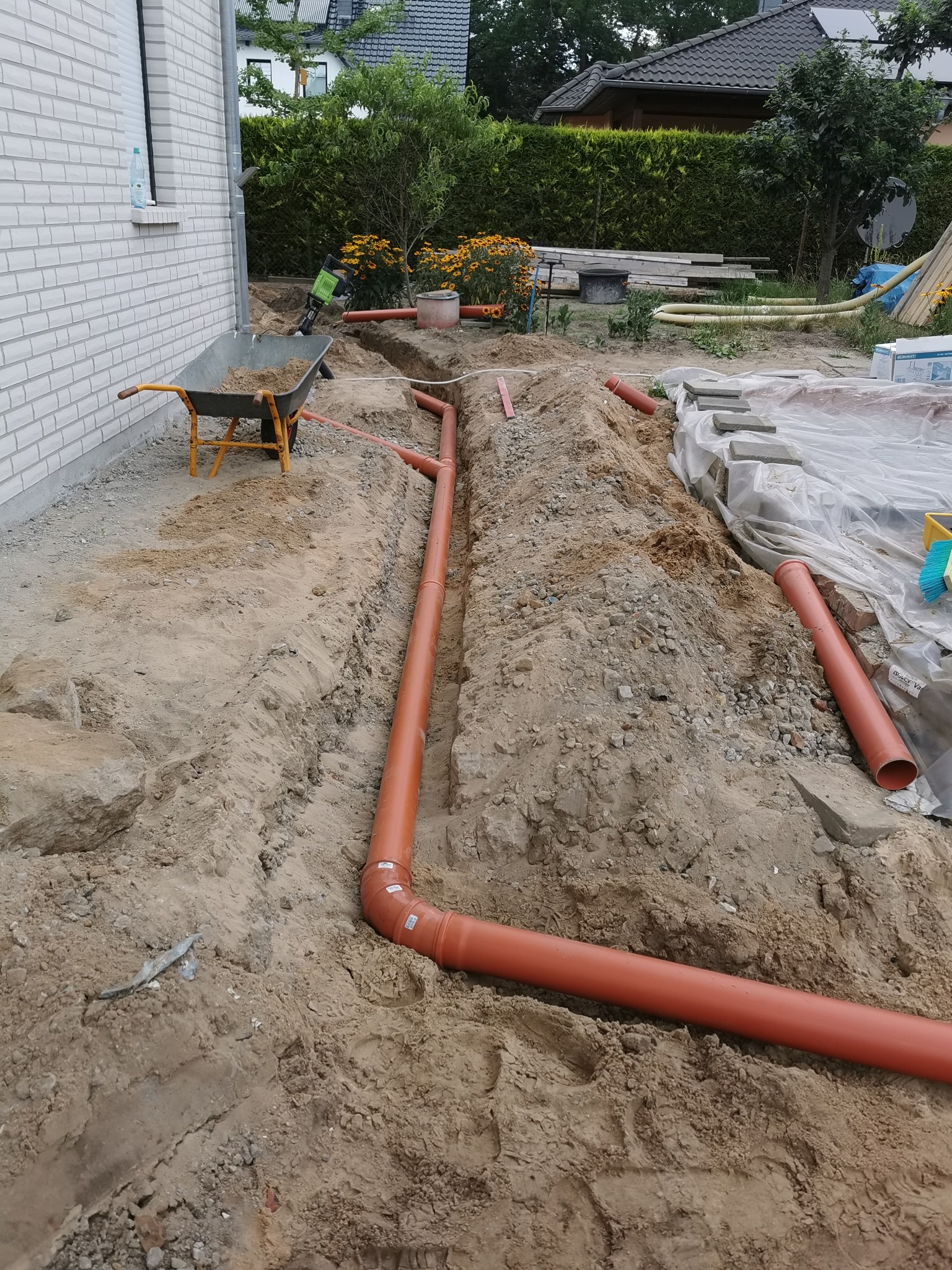 Pomarańczowe rury kanalizacyjne ułożone w wykopie wzdłuż ściany budynku, taczka z ziemią i narzędzia w tle.