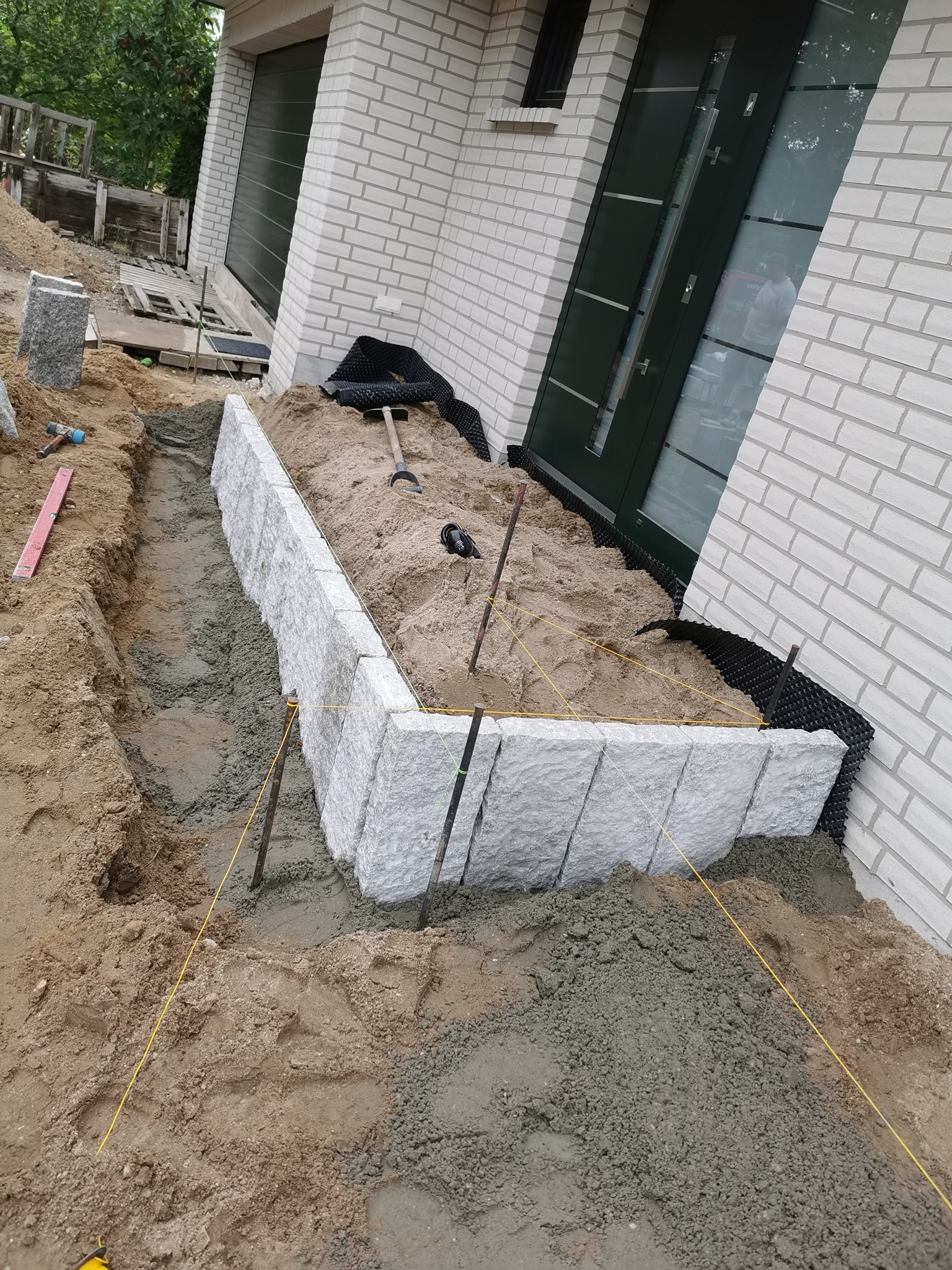 Budowa obrzeża rabaty kwiatowej z kamiennych bloków przy ścianie domu z białej cegły, widoczny wykop, podsypka piaskowa, narzędzia i poziomica.