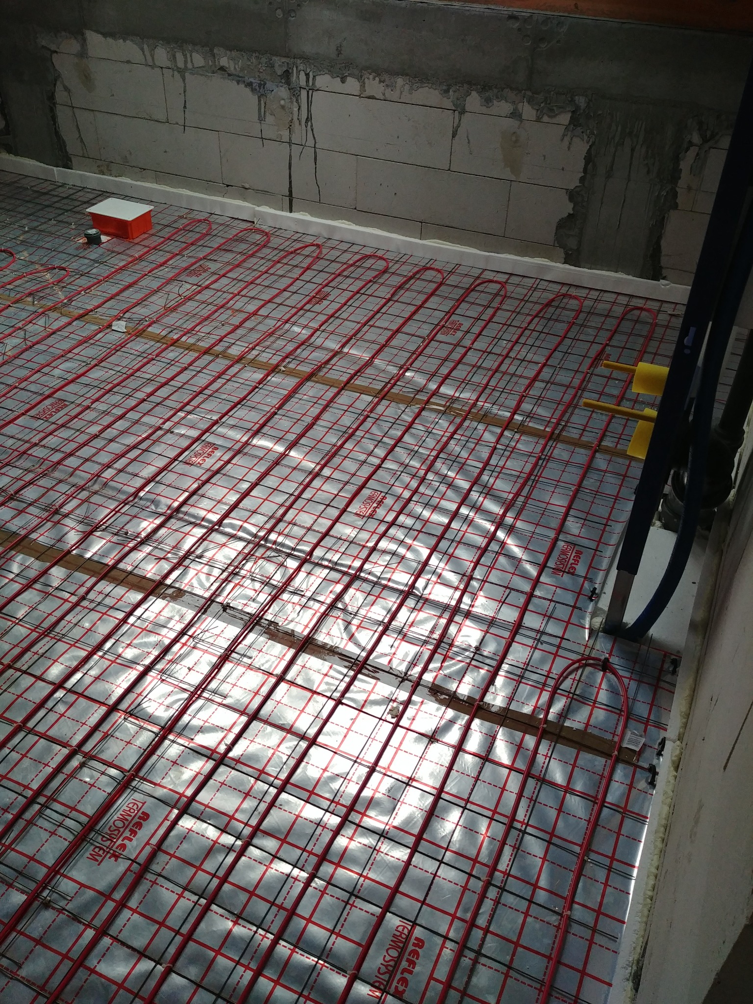 Rozłożona instalacja ogrzewania podłogowego z czerwonymi rurkami na srebrnej folii refleksyjnej, widoczne drewniane listwy mocujące i fragment surowej ściany z bloczków betonowych.