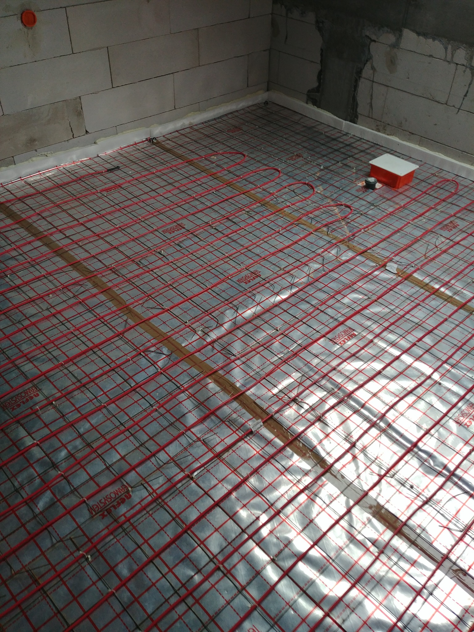 Czerwone kable grzewcze ułożone w siatkę na srebrnej folii izolacyjnej, przygotowane do zalania wylewką w surowym pomieszczeniu z bloczków.