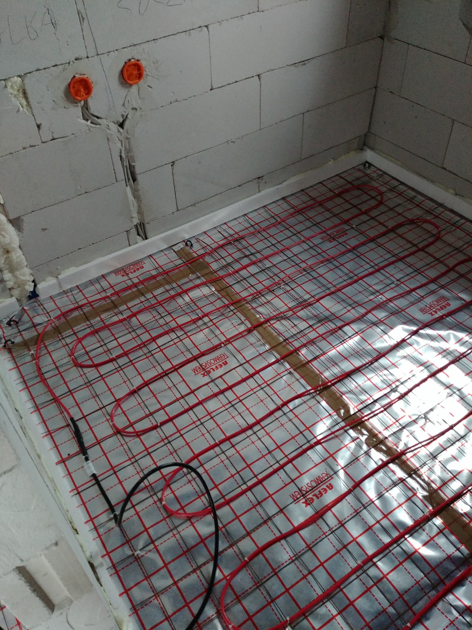 Montaż ogrzewania podłogowego: czerwone kable grzewcze Reflex Termosystem ułożone na srebrnej folii izolacyjnej, widoczne fragmenty ściany z betonu komórkowego i puszki elektryczne.