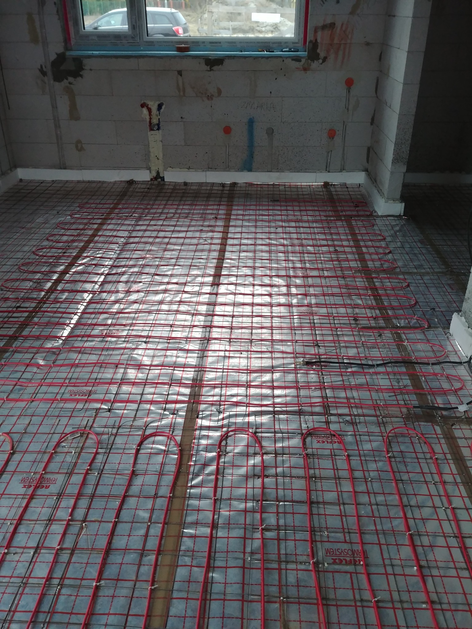 Układanie czerwonych rurek ogrzewania podłogowego na srebrnej folii izolacyjnej, widoczne szare ściany z pustaków i wystające rury instalacji.