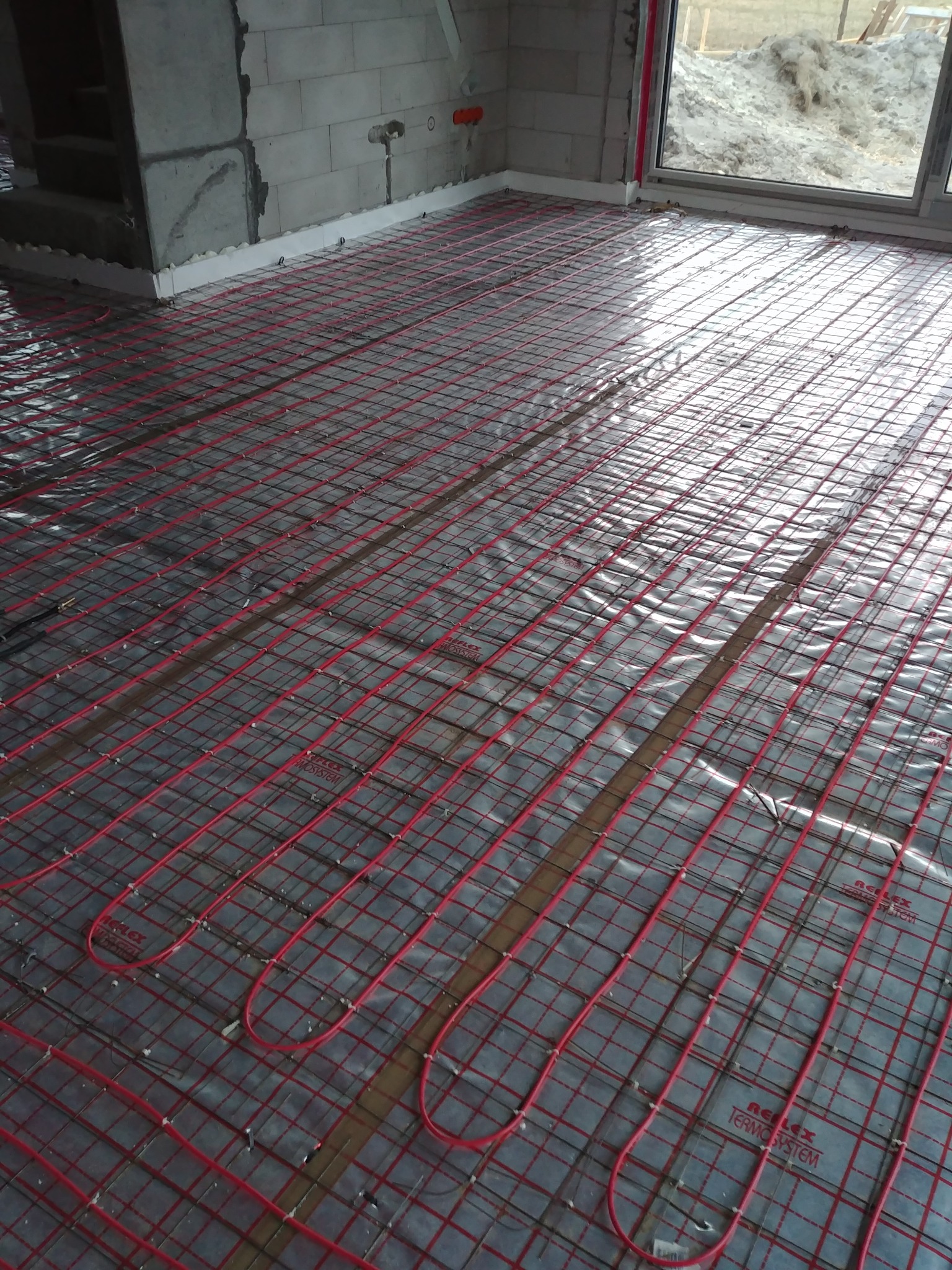 Rozłożona instalacja ogrzewania podłogowego z czerwonymi rurkami na srebrnej folii refleksyjnej w surowym wnętrzu, widoczne bloczki betonowe i okno z widokiem na zewnątrz.