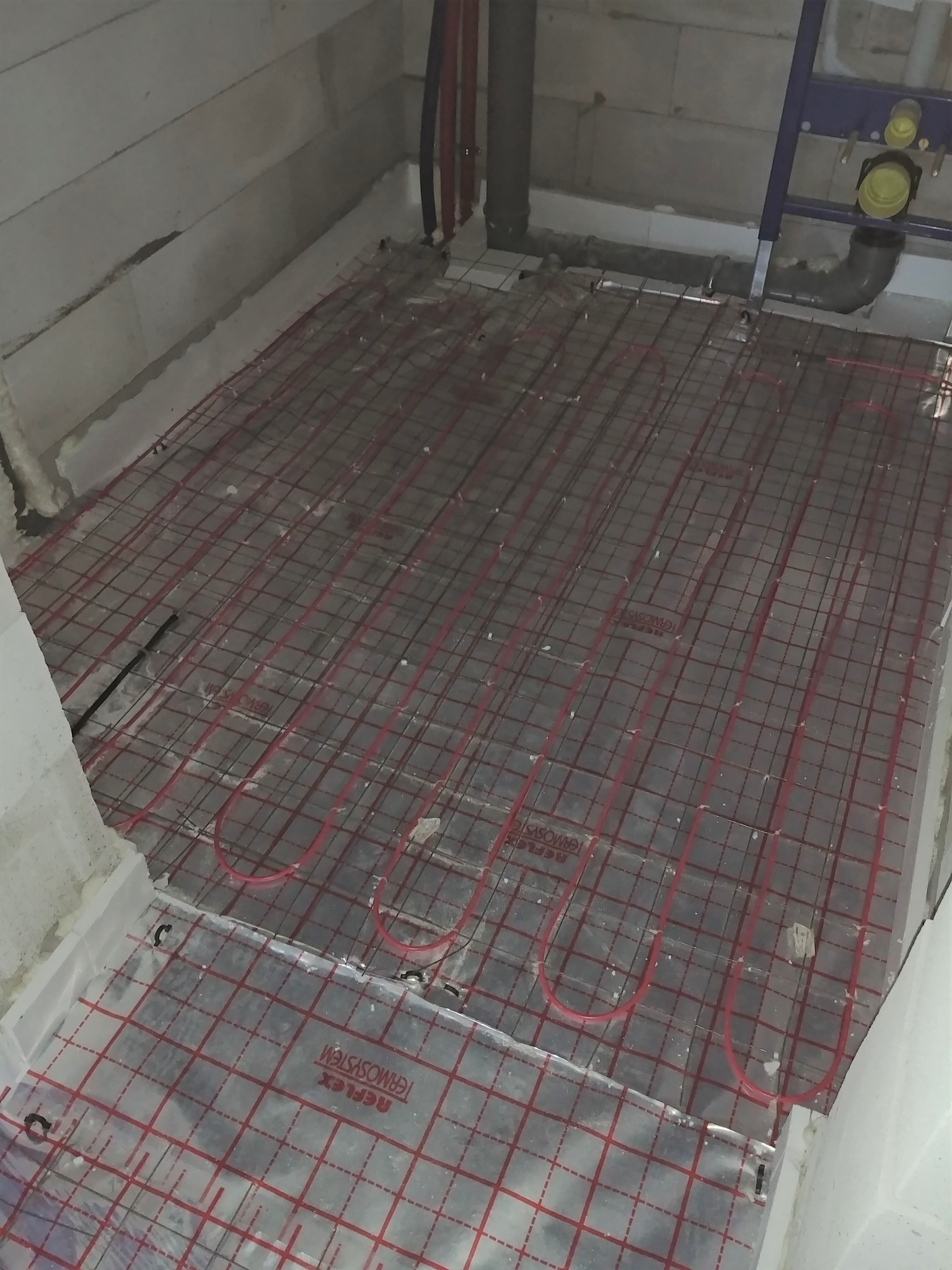 Montaż ogrzewania podłogowego: widok z góry na rozłożone czerwone rury grzewcze na folii z nadrukowaną czerwoną siatką, przygotowane do zalania wylewką, widoczne fragmenty ścian w surowym stanie.