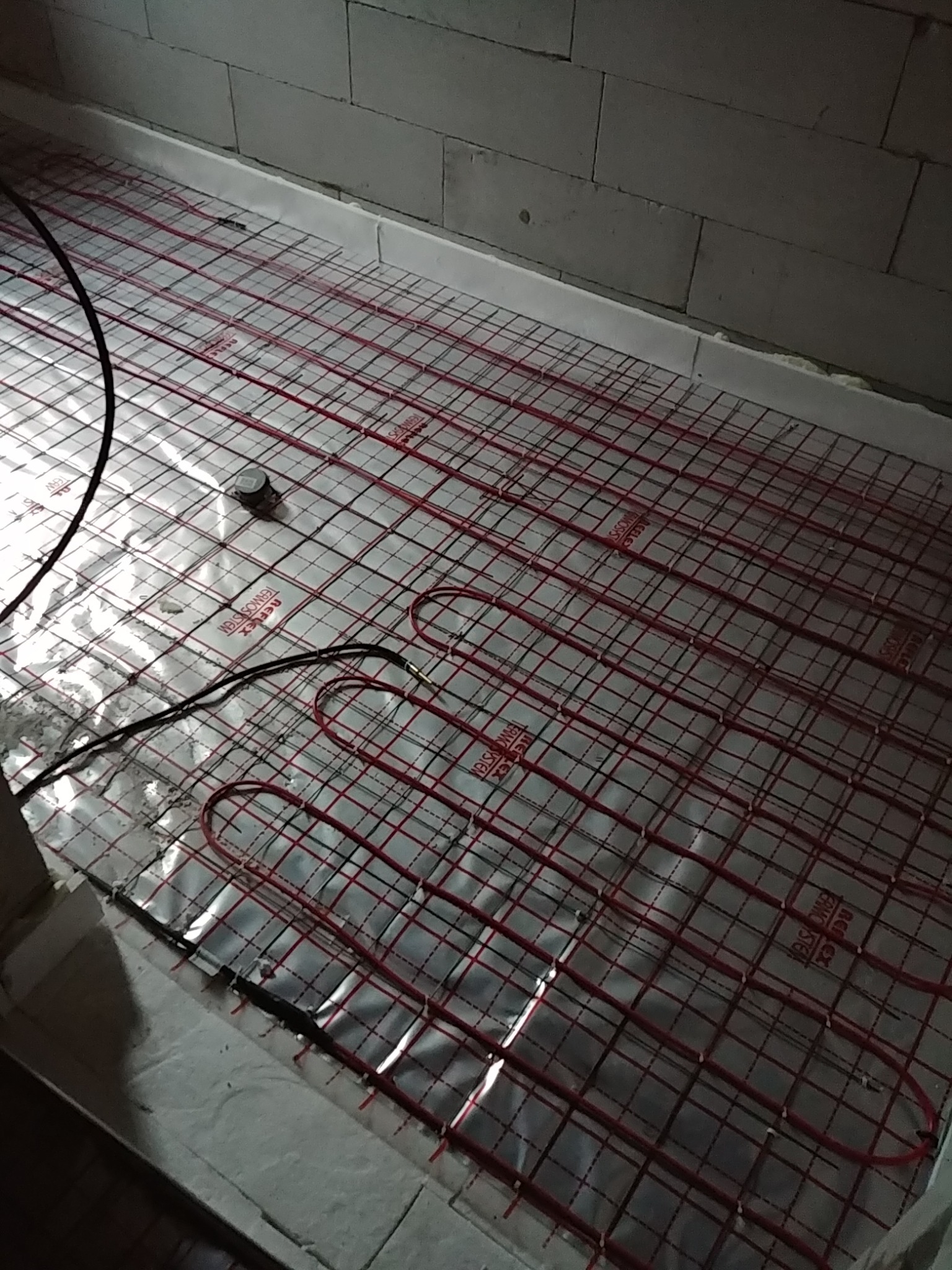 Rozłożona mata grzewcza z czerwonymi przewodami na srebrnej folii, widoczny termostat i przewody zasilające, przygotowana do zalania wylewką.