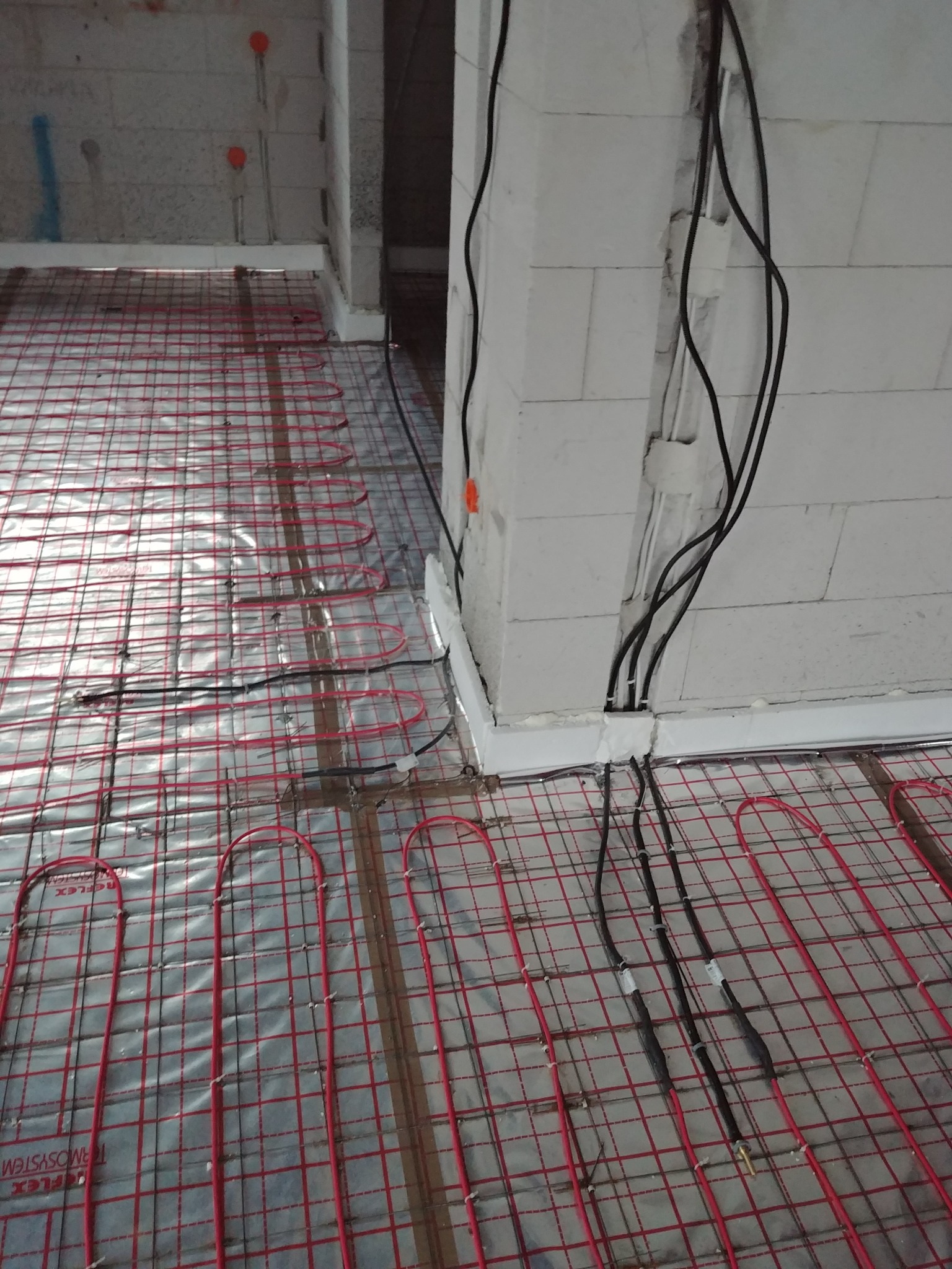 Czerwone rurki ogrzewania podłogowego ułożone na srebrnej folii z widocznymi przewodami elektrycznymi wychodzącymi ze ściany z betonu komórkowego.