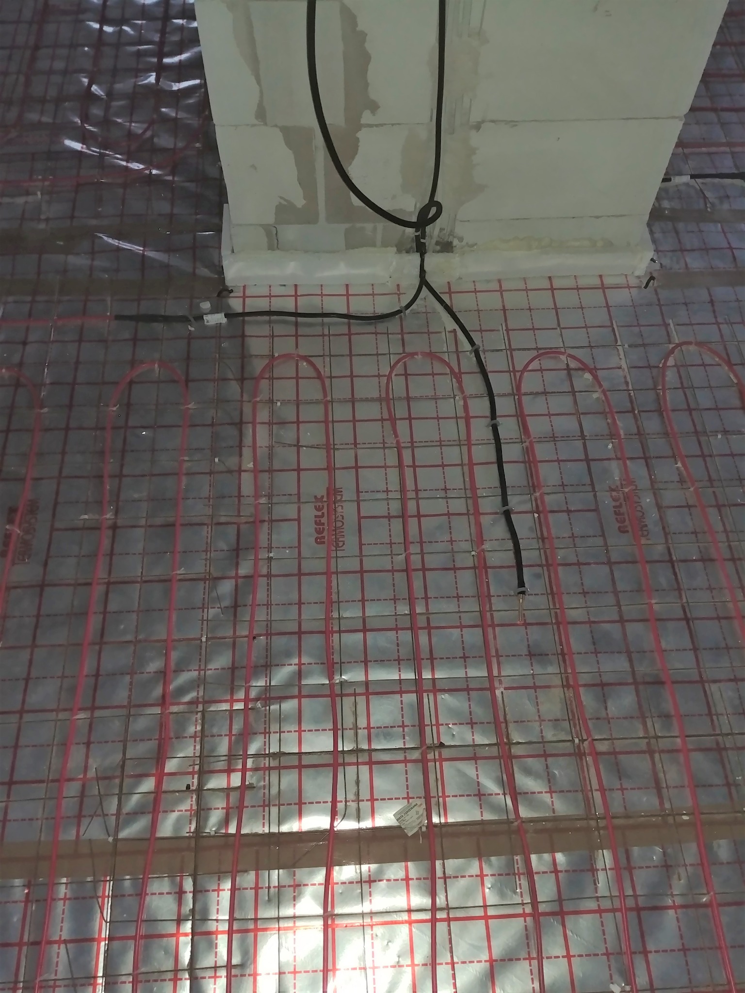 Czerwone kable grzewcze ułożone na folii refleksyjnej, przygotowane do zalania wylewką pod ogrzewanie podłogowe, widoczne czarne przewody zasilające.