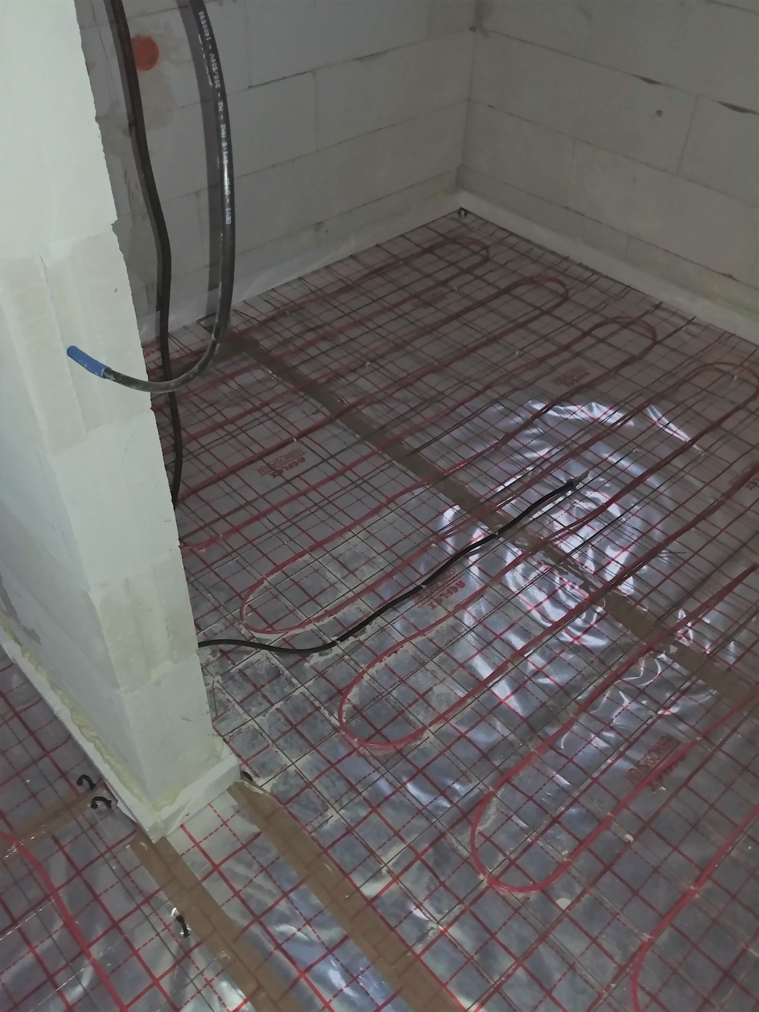 Przygotowanie do instalacji ogrzewania podłogowego: widok na rozłożoną siatkę grzewczą z przewodami na folii izolacyjnej w surowym pomieszczeniu z białymi ścianami z betonu komórkowego.