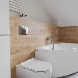 Nowoczesna łazienka na poddaszu z wanną narożną, podwieszaną toaletą i grzejnikiem drabinkowym, wykończona płytkami imitującymi drewno.