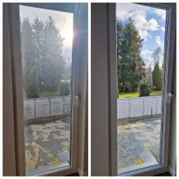 Okno dwudzielne, po lewej stronie brudne, po prawej czyste, widok na podwórko z kostki brukowej, ogrodzenie i drzewa.