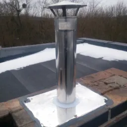 Błyszczący, stalowy komin z daszkiem, zamontowany na dachu pokrytym papą z resztkami śniegu, uszczelniony srebrną blachą i czarnym silikonem, na tle zimowego krajobrazu z drzewami bez liści.