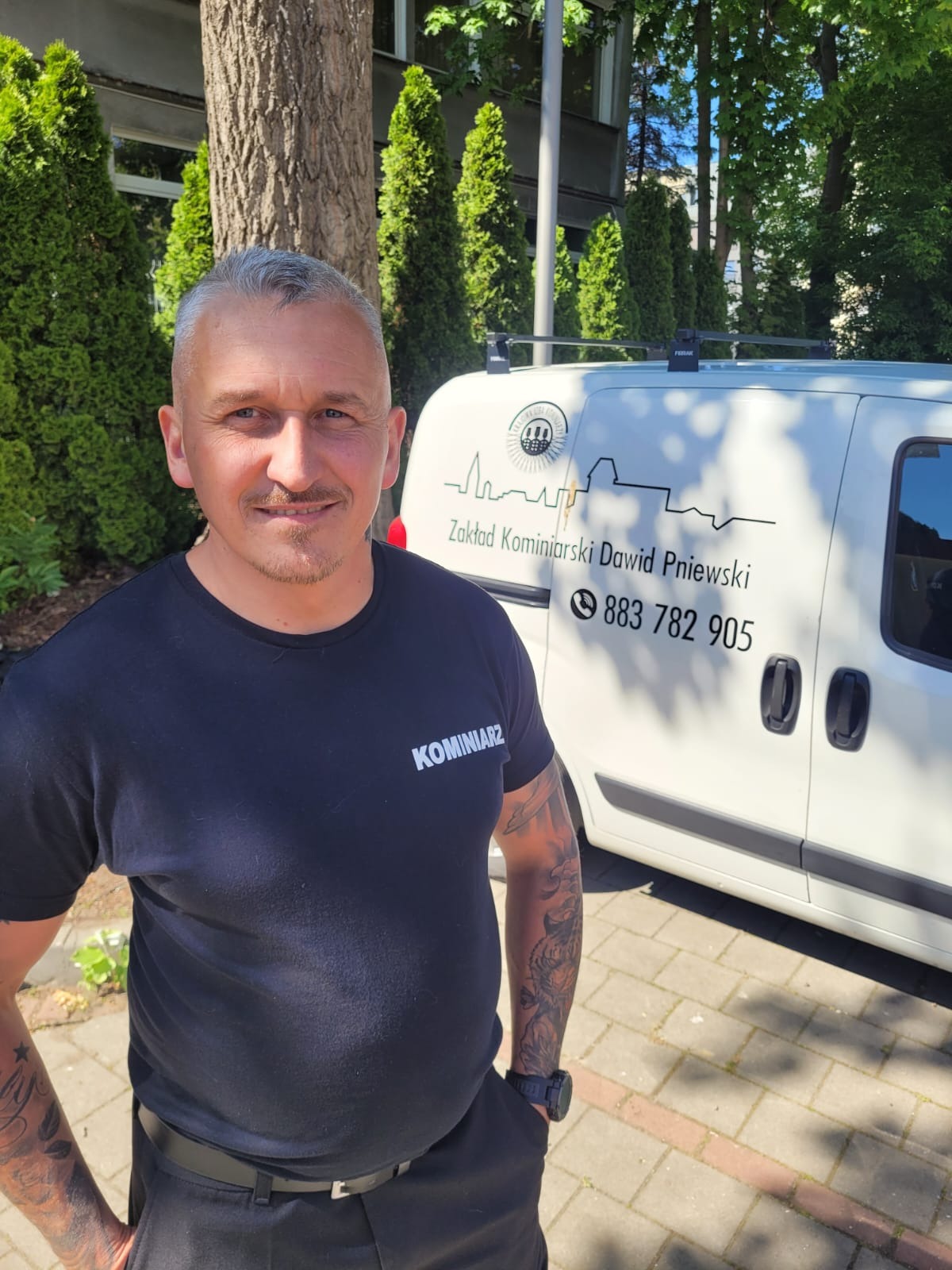 Portret mężczyzny w czarnym T-shircie z napisem 'KOMINIARZ', stojącego na tle białego samochodu firmowego z logo i danymi kontaktowymi Zakładu Kominiarskiego Dawid Pniewski.
